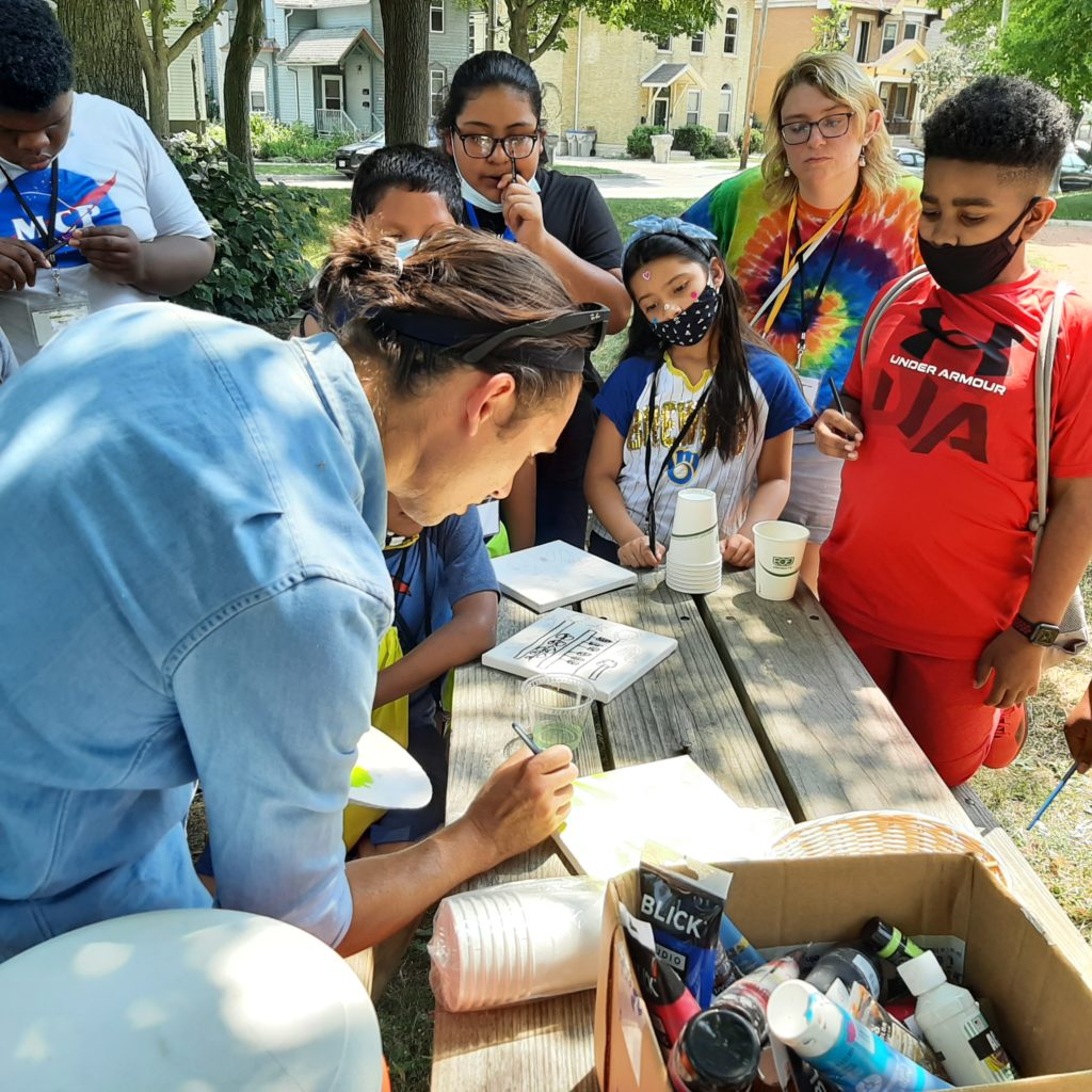 Students gather around a picnic table to watch an artist give a painting demonstration